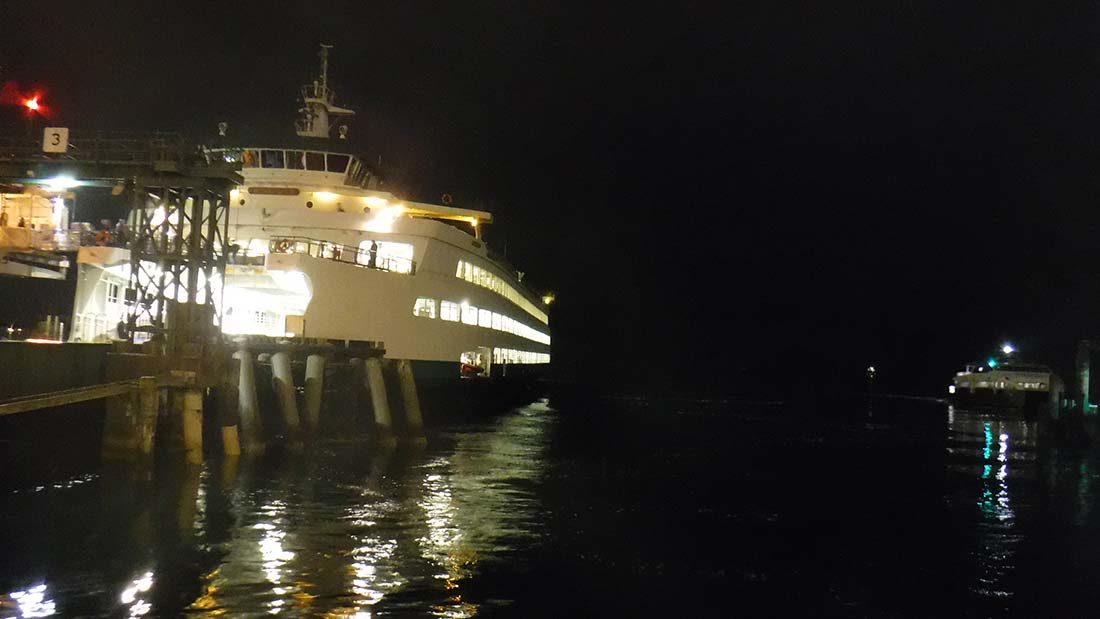 It is night and the sky appears black over the water which is also very dark. There is a ferry at a dock, fully lit and reflecting off the water in the left of the frame. To the right, in the distance, is another ferry.