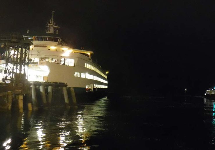 It is night and the sky appears black over the water which is also very dark. There is a ferry at a dock, fully lit and reflecting off the water in the left of the frame. To the right, in the distance, is another ferry.