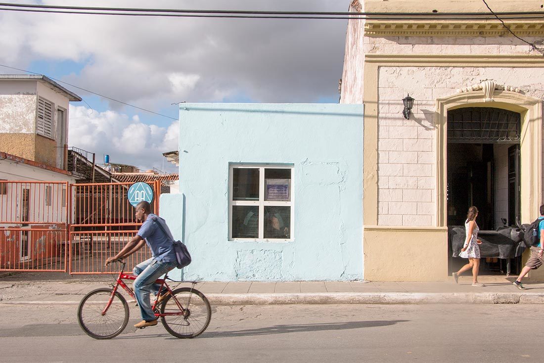 This photograph of a street shows a red fence, a light blue building with a four pane windows, and a beige building with a black lamp. A man rides a bike with a bag hanging down his back on a red bicycle. On the sidewalk in front of the beige building is a woman with brown hair wearing a white patterned sleeveless dress.