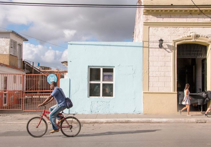 This photograph of a street shows a red fence, a light blue building with a four pane windows, and a beige building with a black lamp. A man rides a bike with a bag hanging down his back on a red bicycle. On the sidewalk in front of the beige building is a woman with brown hair wearing a white patterned sleeveless dress.