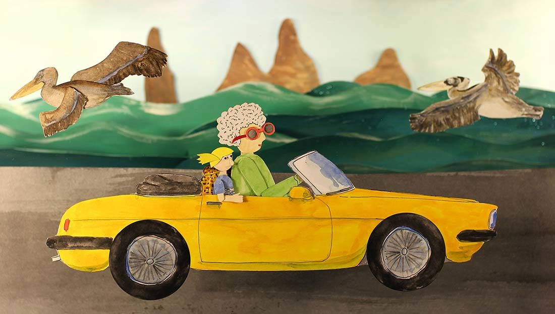 A yellow car drives on a road with waves, sea birds, and tall rocks against a light blue sky behind it. The car is two-door with the top down and animal print seat cover. A woman with white curly hair, a a green shirt, and red sunglasses with drives the car. In the passenger seat is a child with a blue shirt and yellow hair. The image is made out of cut paper.