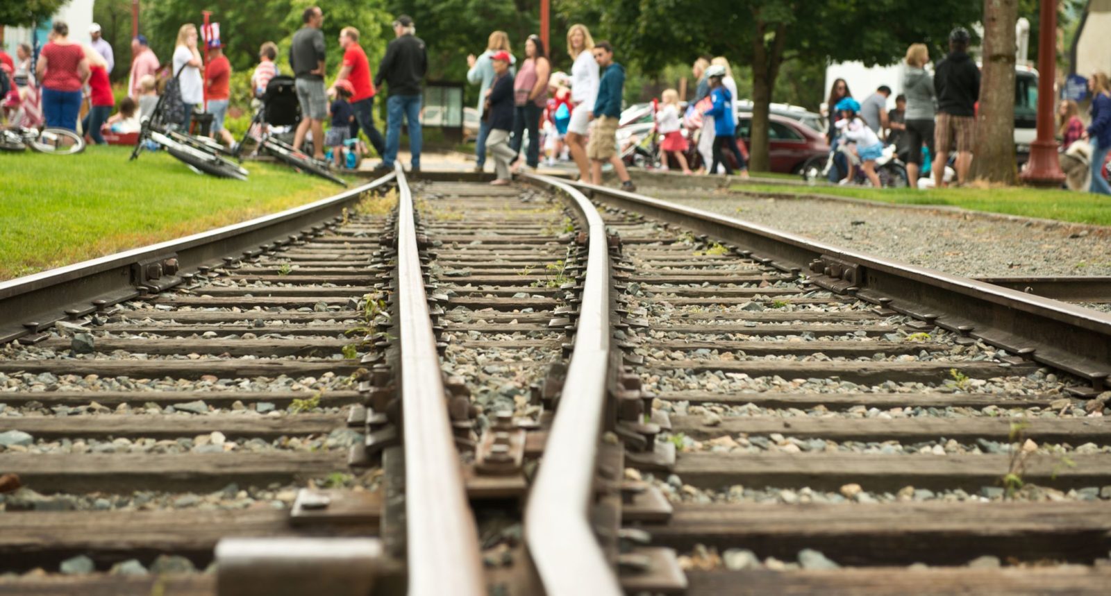 Photographed train tracks emerge from the foreground and extend through the frame. In the distance, children and adults walk across them, with bicycles laying on the ground to the left.