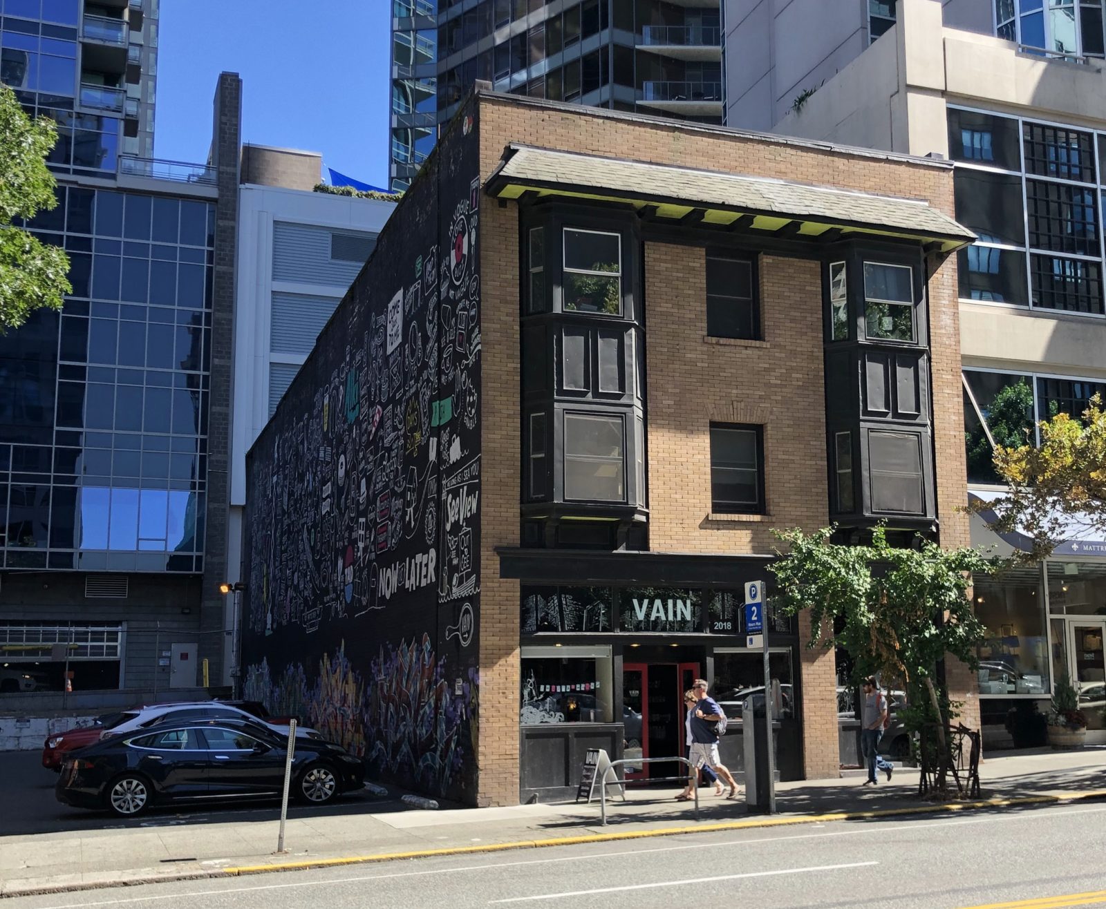 A 3-story, brick building is photographed from across the street. The building has bay windows on the 2nd and 3rd floors and a storefront with a sign reading "VAIN" on the ground floor. The building is surrounded by high rise buildings with glass windows.