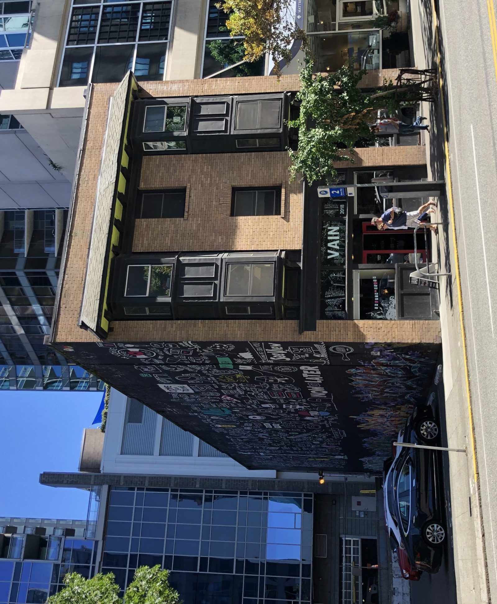 A 3-story, brick building is photographed from across the street. The building has bay windows on the 2nd and 3rd floors and a storefront with a sign reading "VAIN" on the ground floor. The building is surrounded by high rise buildings with glass windows.