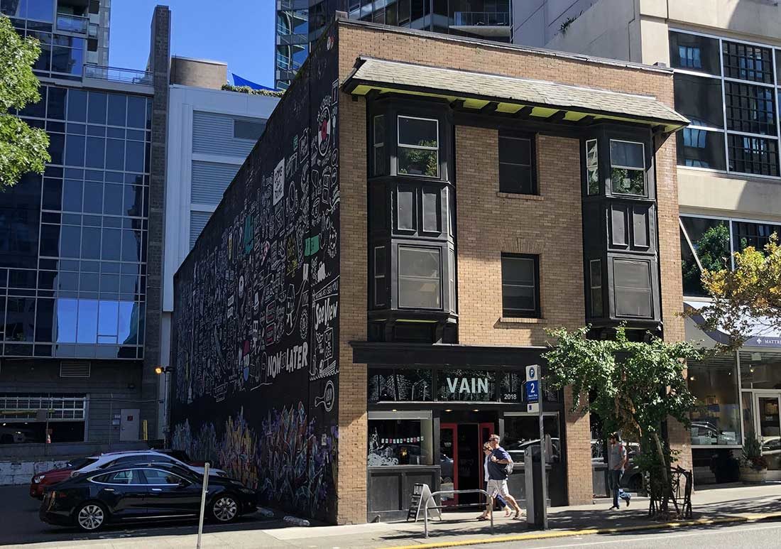 A 3-story, brick building is photographed from across the street. The building has bay windows on the 2nd and 3rd floors and a storefront with a sign reading "VAIN" on the ground floor. The building is surrounded by high rise buildings with glass windows.