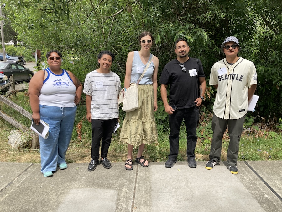 Five people stand on a sidewalk in front of green trees, smiling at the camera. They are casually dressed, each holding papers, and appear to be outdoors on a sunny day.