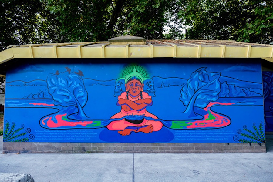 A vibrant mural shows a meditative figure sitting cross-legged with an orange body and green headdress, surrounded by blue leaves, pink and green streams, with trees and a river in the background.