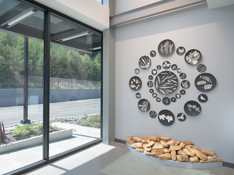 Wall art featuring black circular panels with white botanical and insect designs is arranged in a spiral above a display of tan stones, next to large windows overlooking a road and trees.
