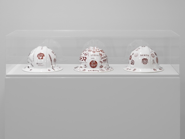 Three white construction helmets decorated with various red logos and text are displayed side by side in a clear case on a white pedestal against a plain white wall.