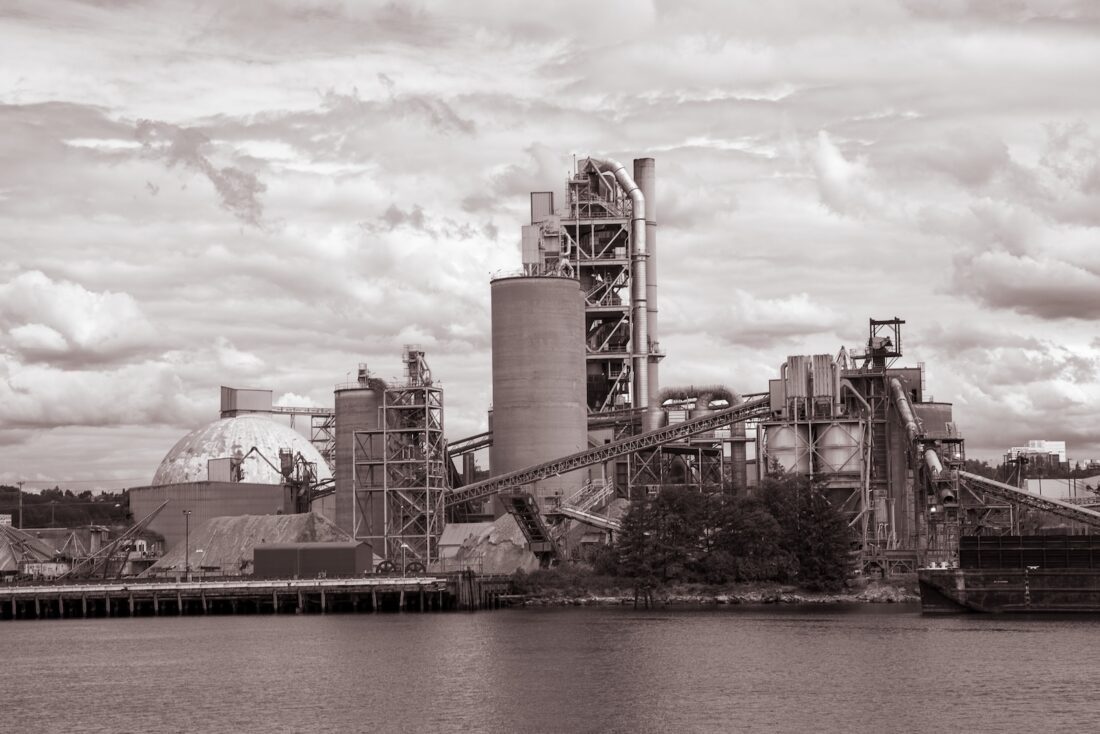 A large industrial factory with multiple silos, towers, and conveyors sits by the waterfront, surrounded by trees and under a cloudy sky. The image is in sepia tone.