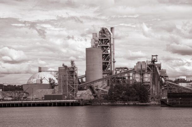 A large industrial factory with multiple silos, towers, and conveyors sits by the waterfront, surrounded by trees and under a cloudy sky. The image is in sepia tone.