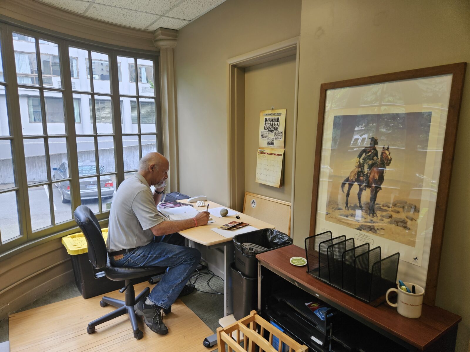A man is sitting at a table reading a document. He is wearing a grey shirt, blue jeans and sneakers. To his left is a large window with many window panes. To his right is a print of a Buffalo Soldier in a landscape. The picture is on a desk leaning against a wall.