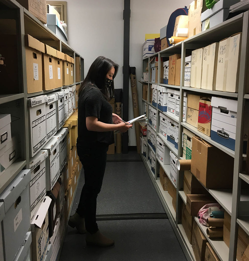 A woman stands between two rows of shelving. She is wearing a black shirt, black pants and beige boots. she is wearing a black mask covering the lower part of her face. The shelves are filled with archival boxes from floor to ceiling. 