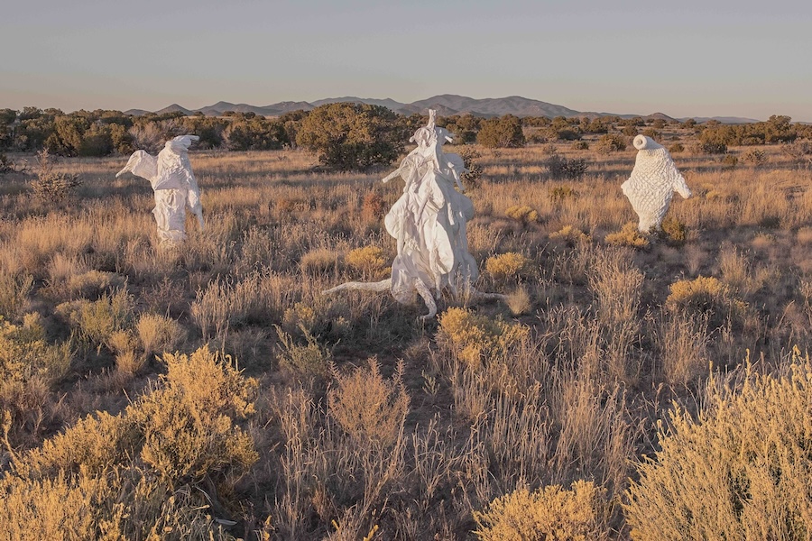 Three figures draped in white, ghost-like fabric stand in a dry, grassy field with bushes, under a clear sky at sunset; distant mountains and trees are visible in the background.