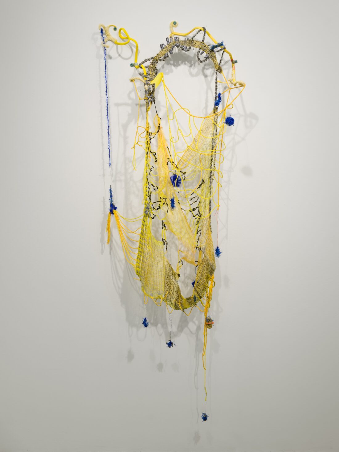 A web-like abstract sculpture made of yellow and blue yarn, string, and mixed materials hangs on a white wall, featuring intricate, netted patterns and organic, looping shapes.