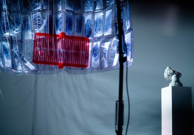 A translucent installation with blue-toned panels and a red central section hangs from above, casting shadows. To the right, a small sculpture sits on a white pedestal under focused lighting.