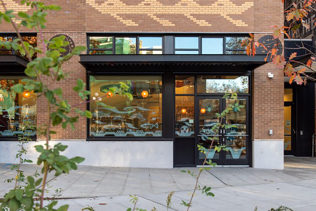 A brick building with large windows, decorative white patterns, and round hanging lights inside. There are green plants in the foreground and a tree with autumn leaves near a paved sidewalk.