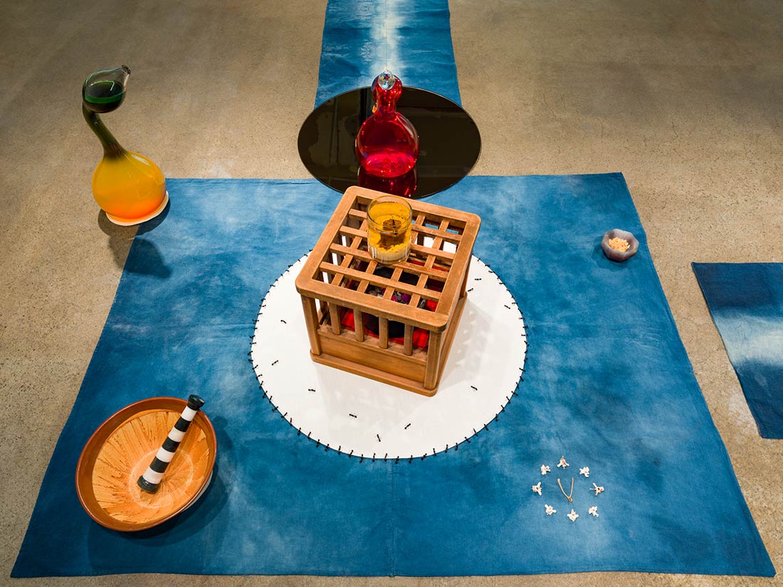 Art installation with an indigo blue cloth featuring a white circle, a wooden cage centerpiece, a yellow glass on top, various ceramic vessels, a red glass bottle, small bowl, and scattered white objects, all on a concrete floor.