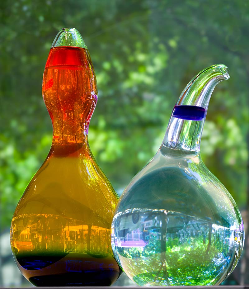Two glass bottles with unique shapes; the left bottle contains layered red, orange, and yellow liquids, while the right bottle is clear with blue liquid bands. Both are set against a blurred green outdoor background.