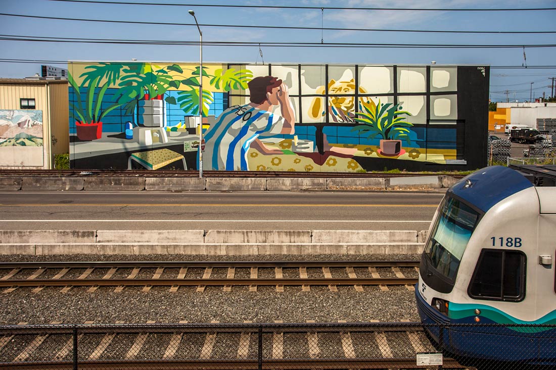 A mural on a building shows a person in a striped shirt sitting at a table, looking out a window with plants inside and a desert scene outside. A train is passing on tracks in the foreground.