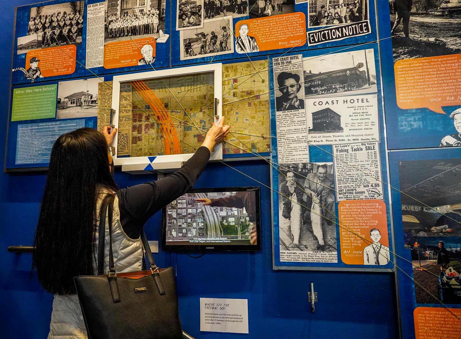A woman examines a display at a museum exhibit featuring historical photos, maps, documents, and eviction notices on a blue wall. She is lifting a framed map.