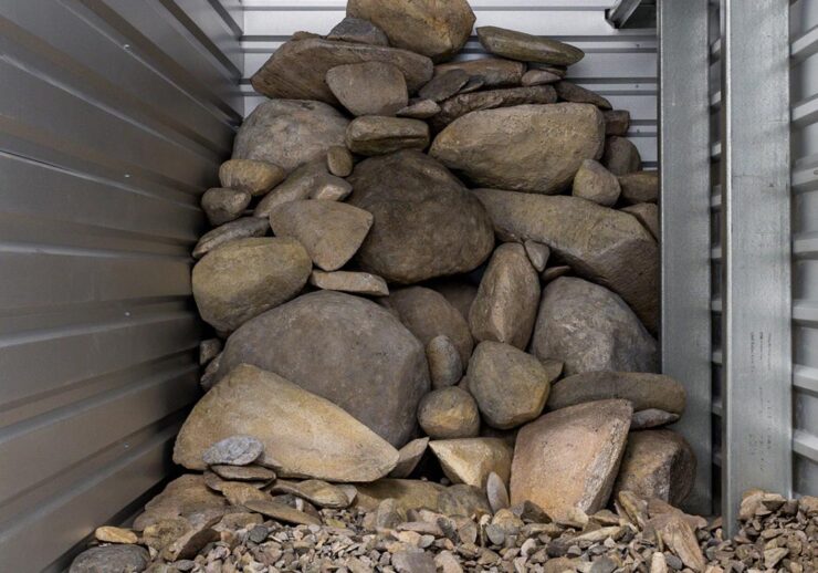 A pile of variously sized rocks and stones stacked in the corner of a metal-walled storage unit, with smaller pebbles scattered on the concrete floor in front.