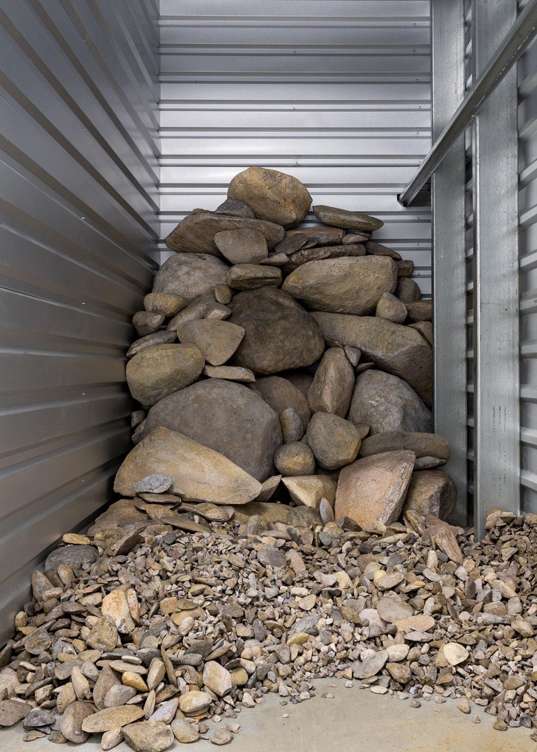 A pile of variously sized rocks and stones stacked in the corner of a metal-walled storage unit, with smaller pebbles scattered on the concrete floor in front.