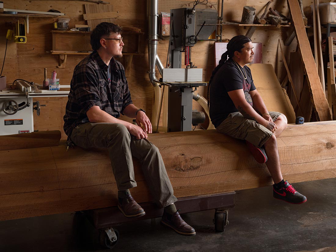 Two people sit side by side on a large wooden beam in a woodshop, surrounded by tools and wooden planks. Both appear thoughtful, looking towards the right, with soft lighting highlighting the workshop environment.