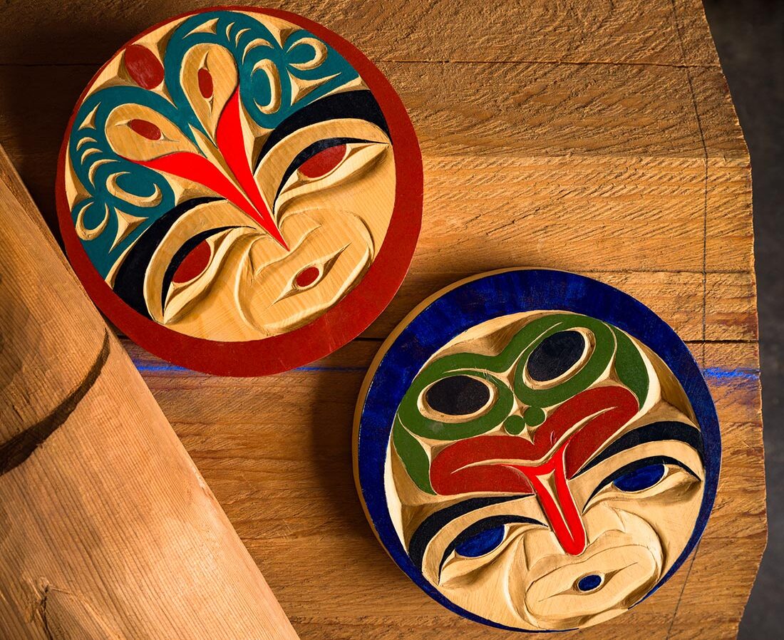 Two round, colorful, intricately carved wooden masks with stylized faces and bold patterns rest on a wooden surface. One mask has red and teal details, the other features green and blue accents.