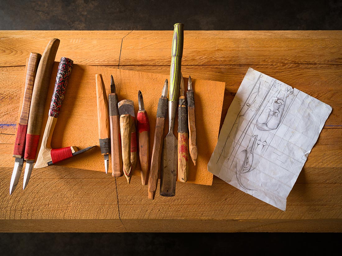 A set of wood carving tools with wooden handles is arranged on a wooden surface beside a piece of orange fabric and a sheet of paper with sketches and notes.