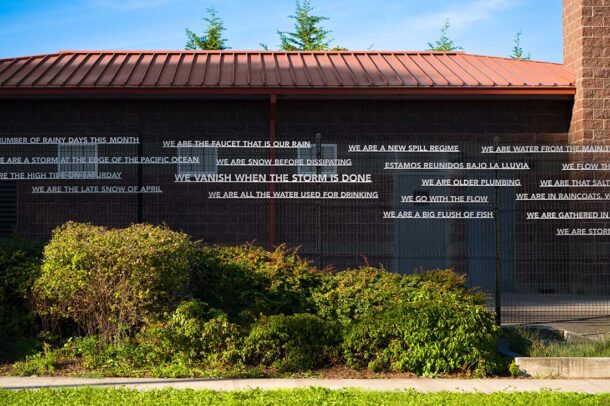 A brick building with metal fencing and greenery in front, displaying poetic phrases in white text strips across the wall, discussing water, rain, and storms. Blue sky and tree tops are visible in the background.