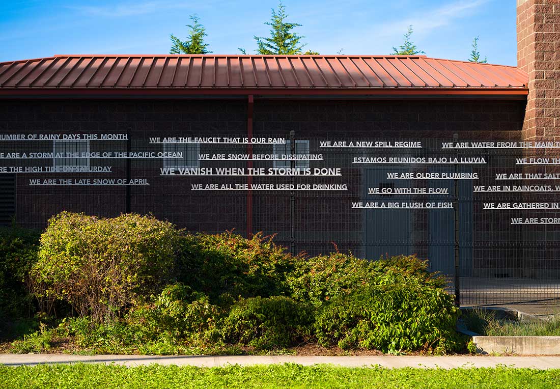 A brick building with metal fencing and greenery in front, displaying poetic phrases in white text strips across the wall, discussing water, rain, and storms. Blue sky and tree tops are visible in the background.