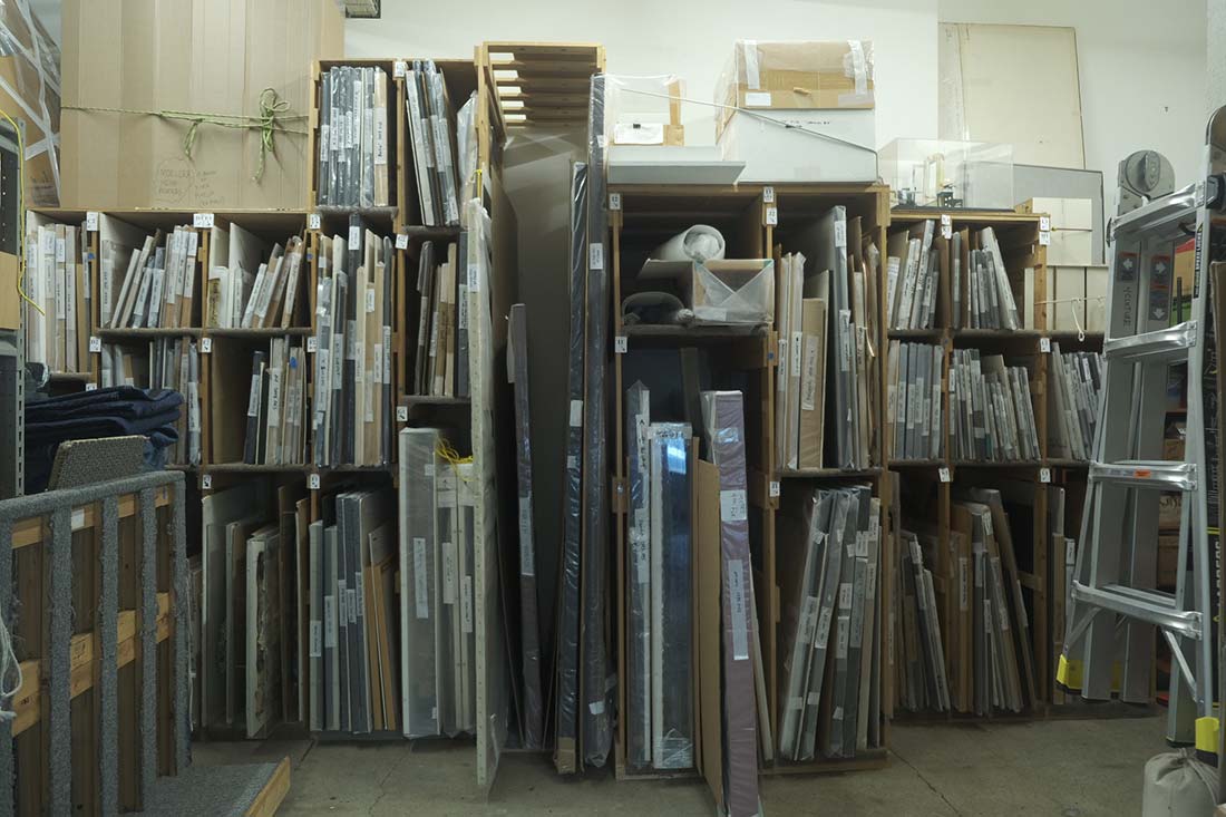 Shelves filled with framed artwork and canvases of various sizes are stacked vertically in a storage room. Cardboard boxes and packing material are visible on the top shelves and floor. A metal ladder stands on the right.