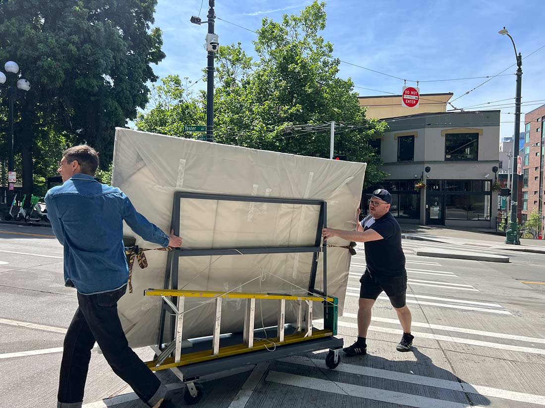 Two men push a large, wrapped object on a wheeled cart across a city street on a sunny day, with trees and buildings in the background.