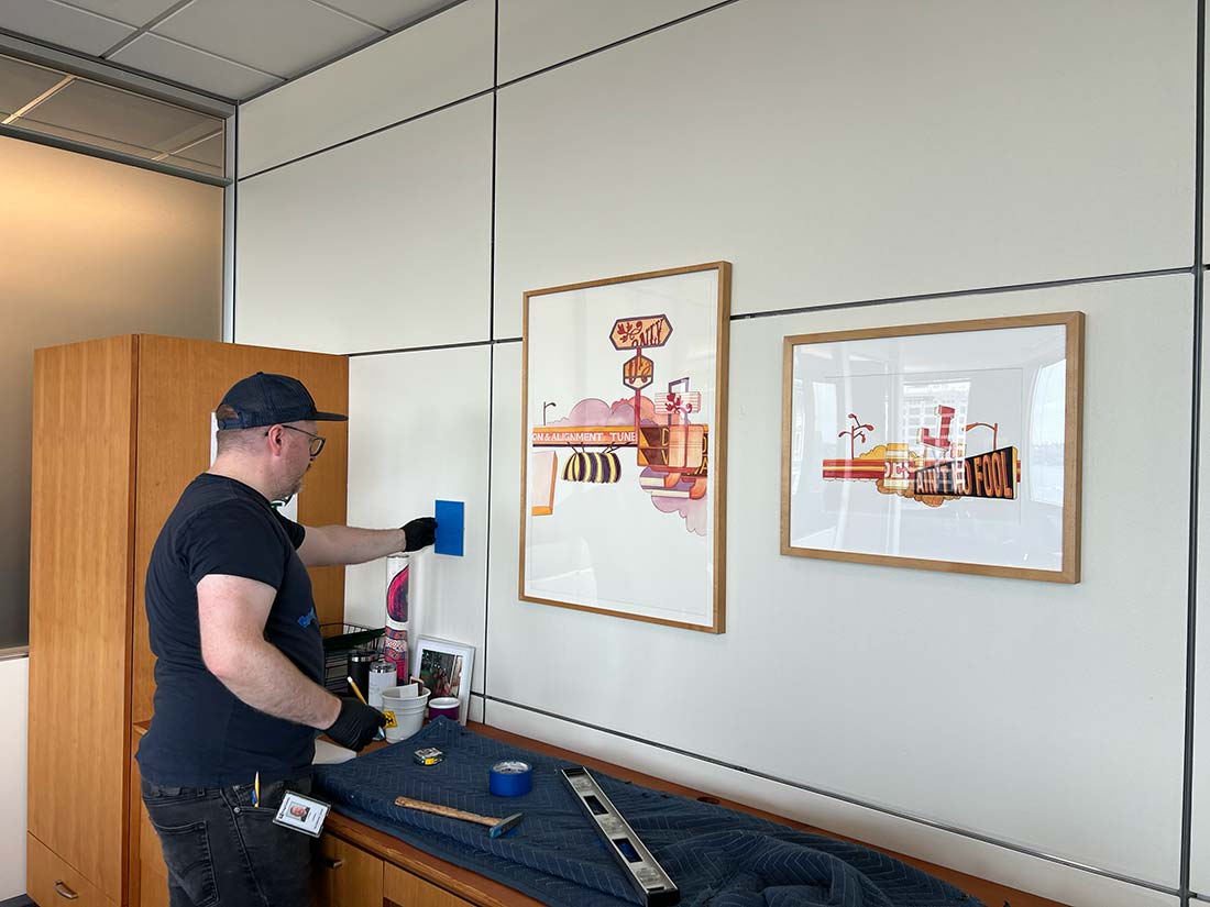 A man wearing a cap and gloves cleans the wall next to two framed artworks in an office, preparing to hang something. Tools and supplies are on a covered cabinet beneath the artwork.