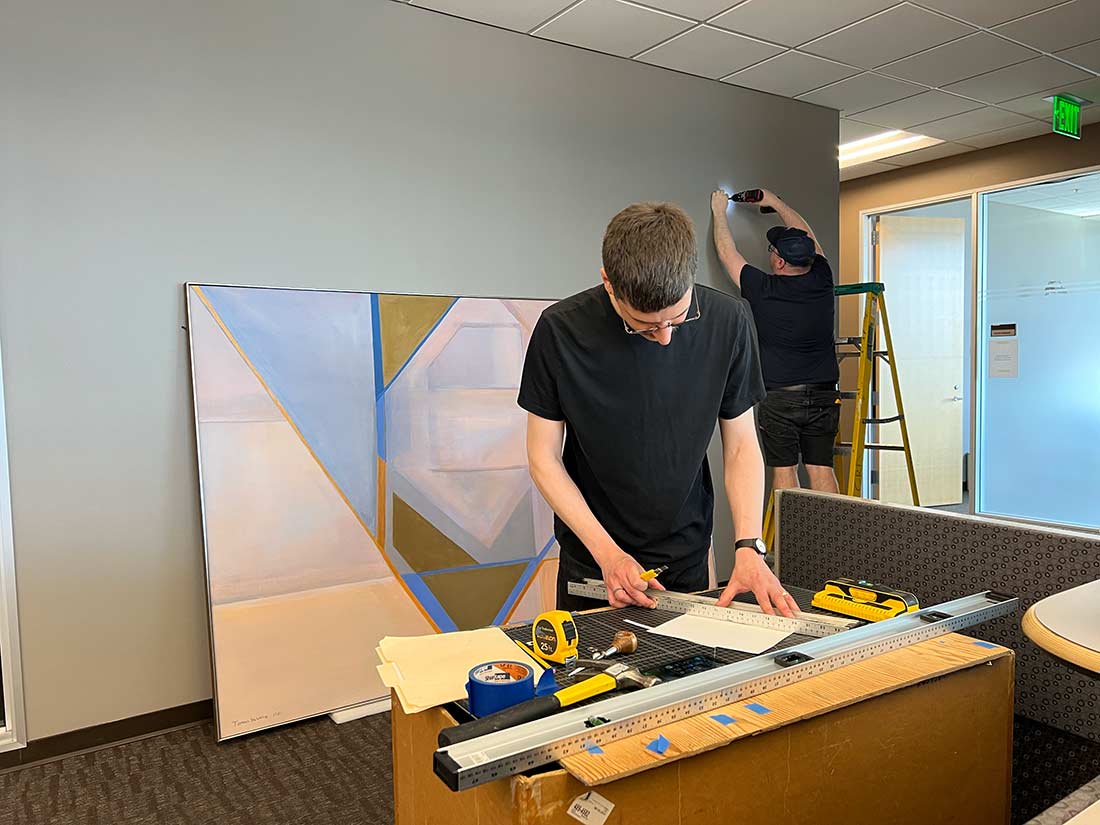 Two men hang artwork in an office. One measures a wall while the other, in the background on a ladder, works near the ceiling. A large abstract painting rests against the wall and tools are spread on a box in the foreground.