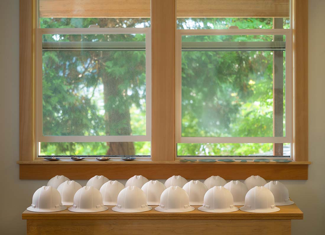 A row of white construction hard hats is lined up on a wooden table in front of a window with greenery visible outside.