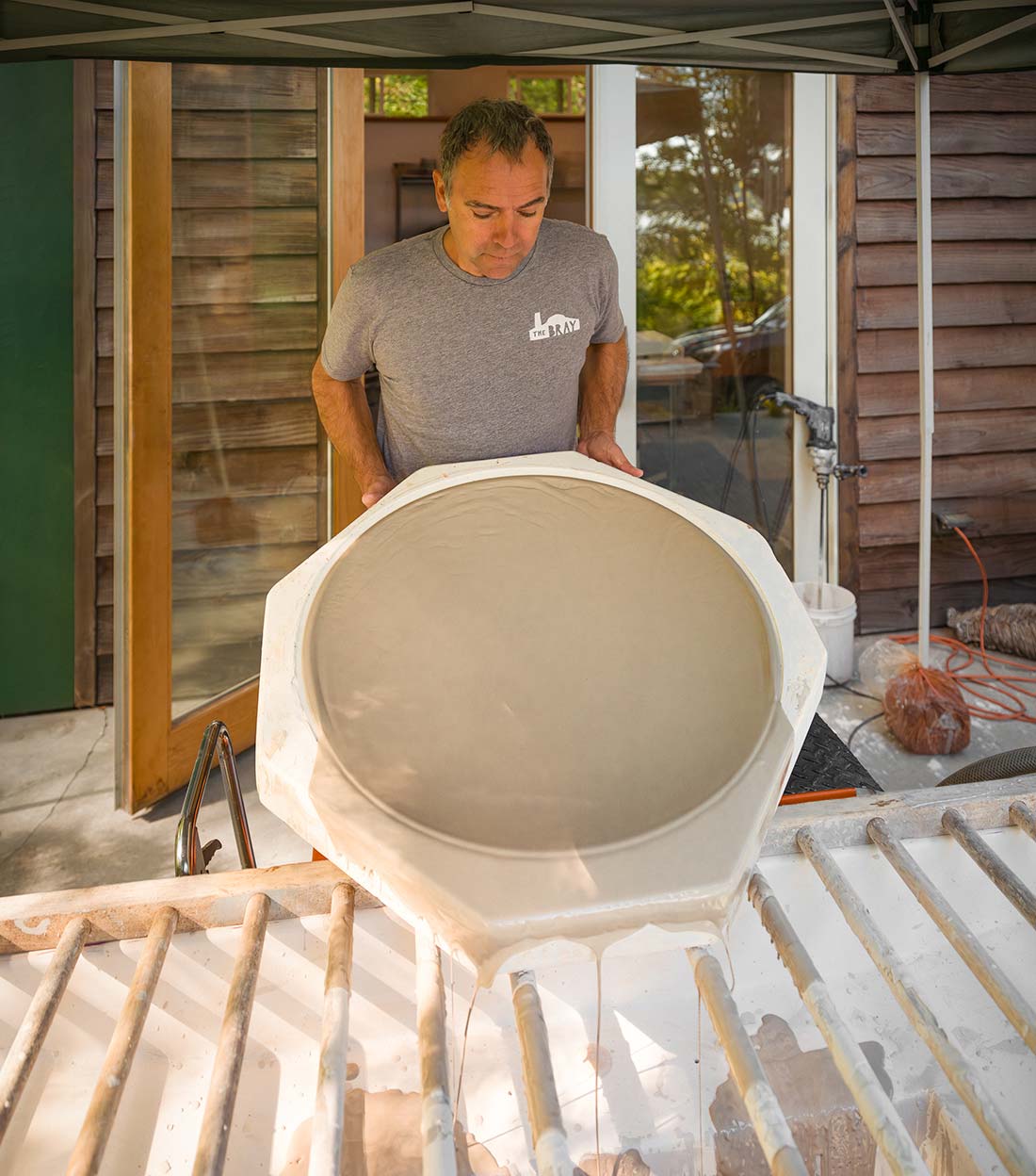 A man in a gray shirt holds a large, circular mold filled with wet cement or plaster, standing outdoors next to a wooden building, with sunlight streaming in from the side.