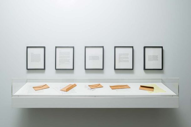 A display case containing envelopes and documents is mounted below five framed texts on a plain white wall in a minimalist gallery setting.