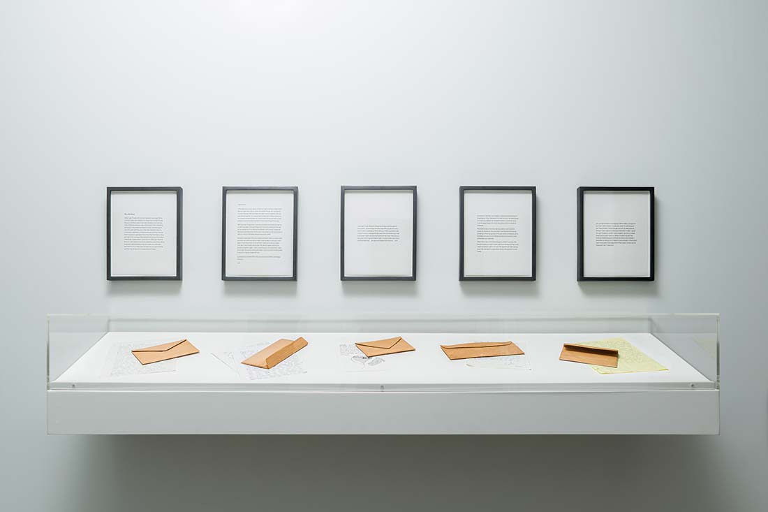 A display case containing envelopes and documents is mounted below five framed texts on a plain white wall in a minimalist gallery setting.