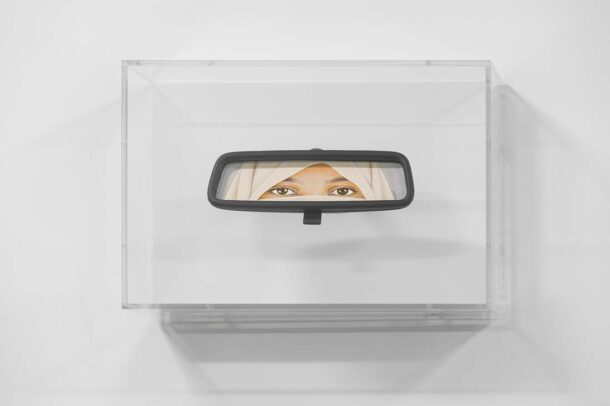 A car rearview mirror mounted on a clear box displays the reflection of a persons eyes wearing a beige headscarf, centered against a plain white background.