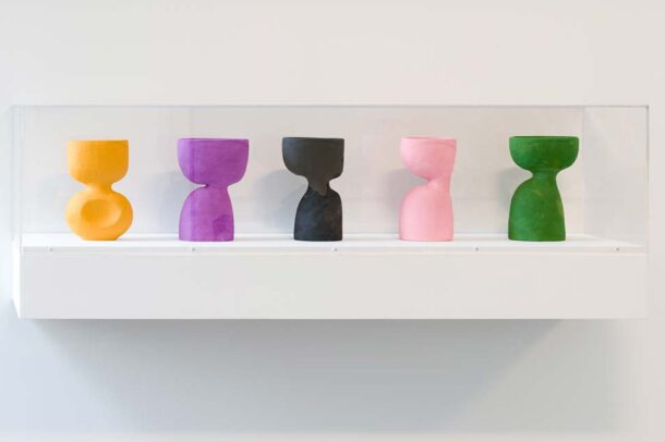Five hourglass-shaped sculptures in orange, purple, black, pink, and green are displayed in a clear case mounted on a white wall.