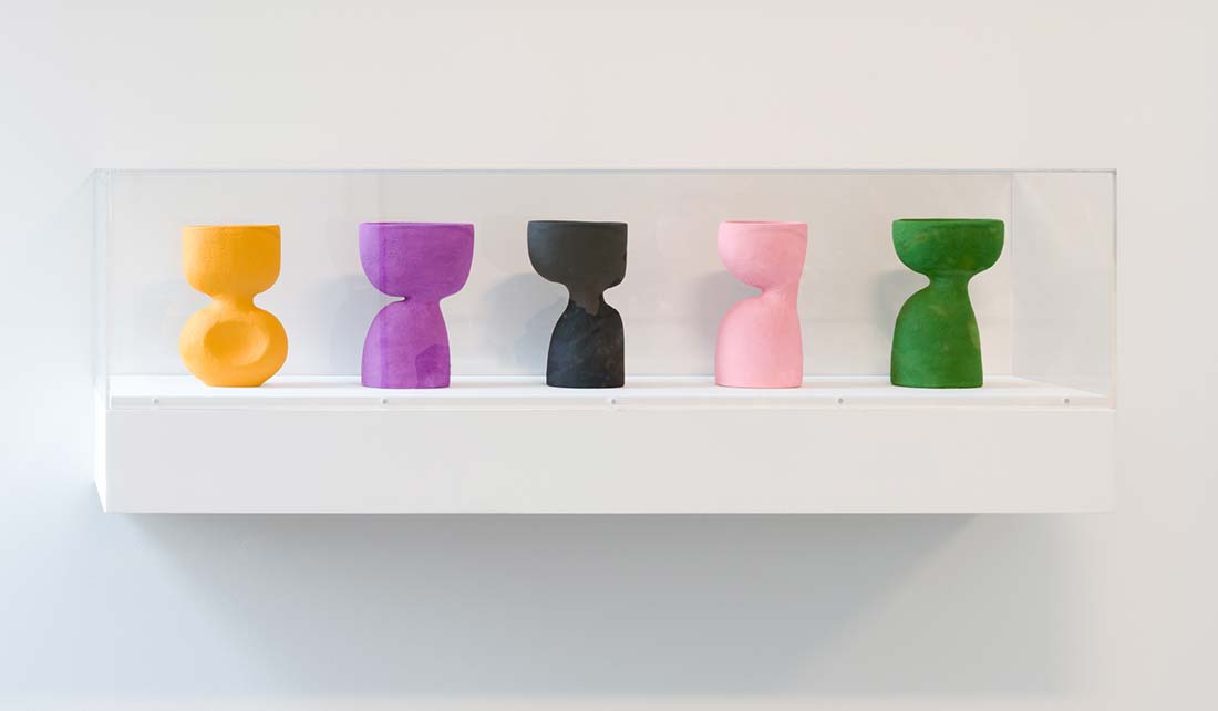 Five hourglass-shaped sculptures in orange, purple, black, pink, and green are displayed in a clear case mounted on a white wall.