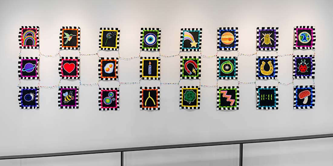 A wall display of 24 colorful, square artworks featuring various bold symbols like hearts, stars, planets, and animals, each bordered in black and white stripes, connected by a string of decorative lights.