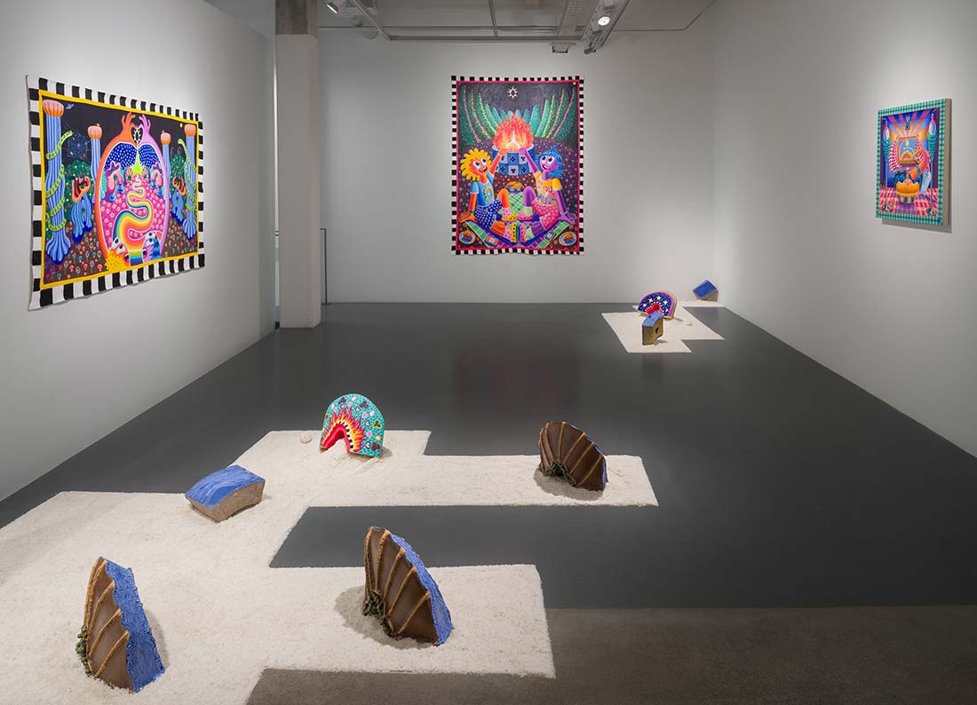 A contemporary art gallery with colorful, whimsical paintings on white walls and vibrant, abstract sculptures displayed on white platforms across a dark floor. The scene is brightly lit and visually dynamic.