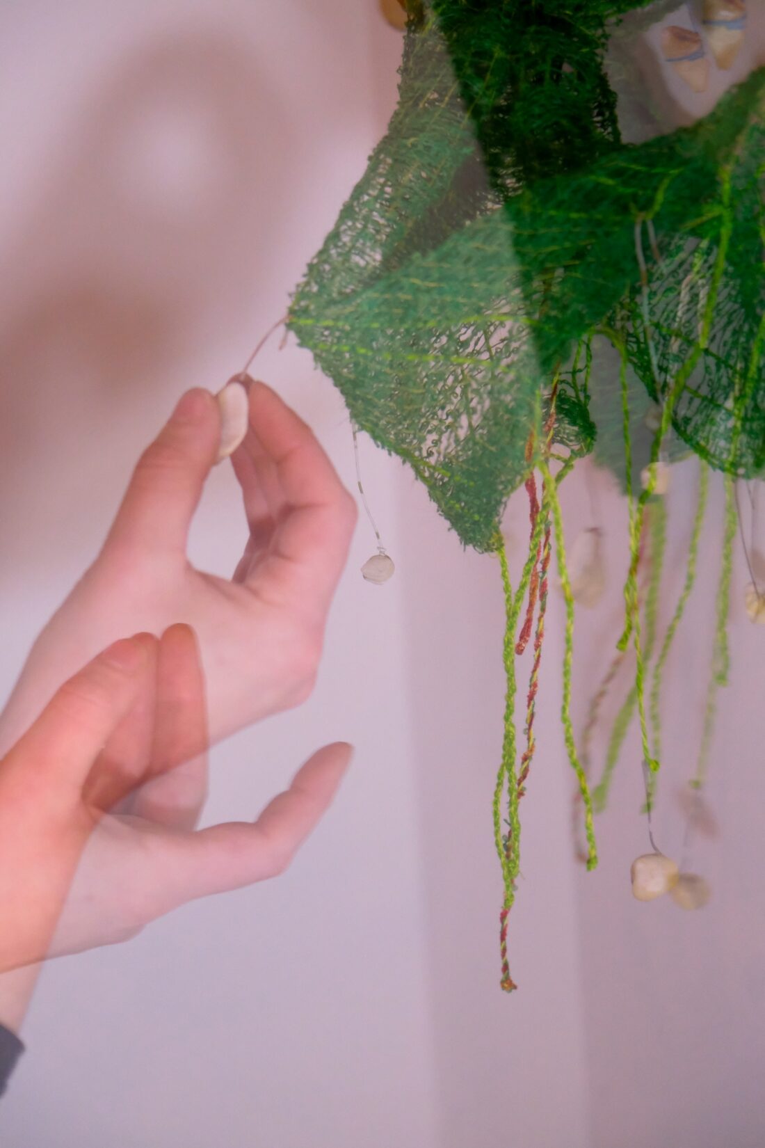 A blurred hand reaches toward hanging objects on green textured threads with a soft, pinkish background. The motion creates a double exposure effect, highlighting the hand’s movement.