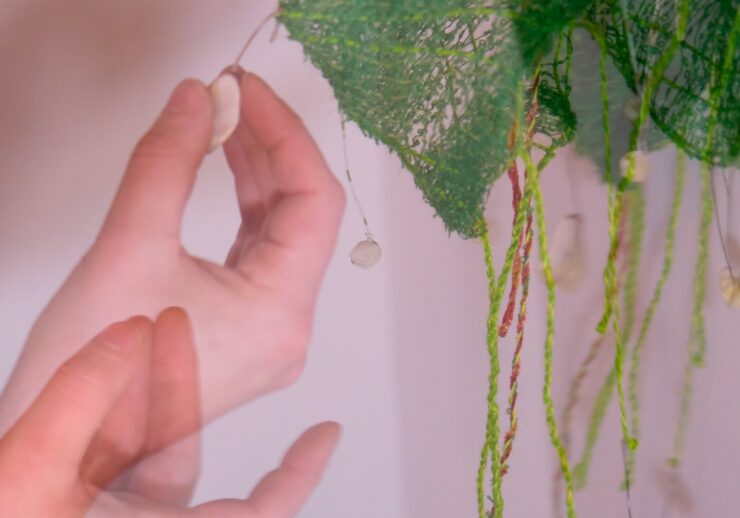 A blurred hand reaches toward hanging objects on green textured threads with a soft, pinkish background. The motion creates a double exposure effect, highlighting the hand’s movement.