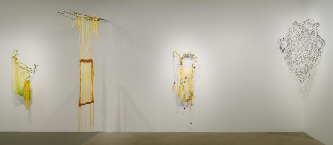 A gallery wall displays four abstract sculptural artworks made from wire, netting, and fibers in yellow, orange, and black, with organic shapes and materials draping or hanging loosely. The floor is smooth concrete.