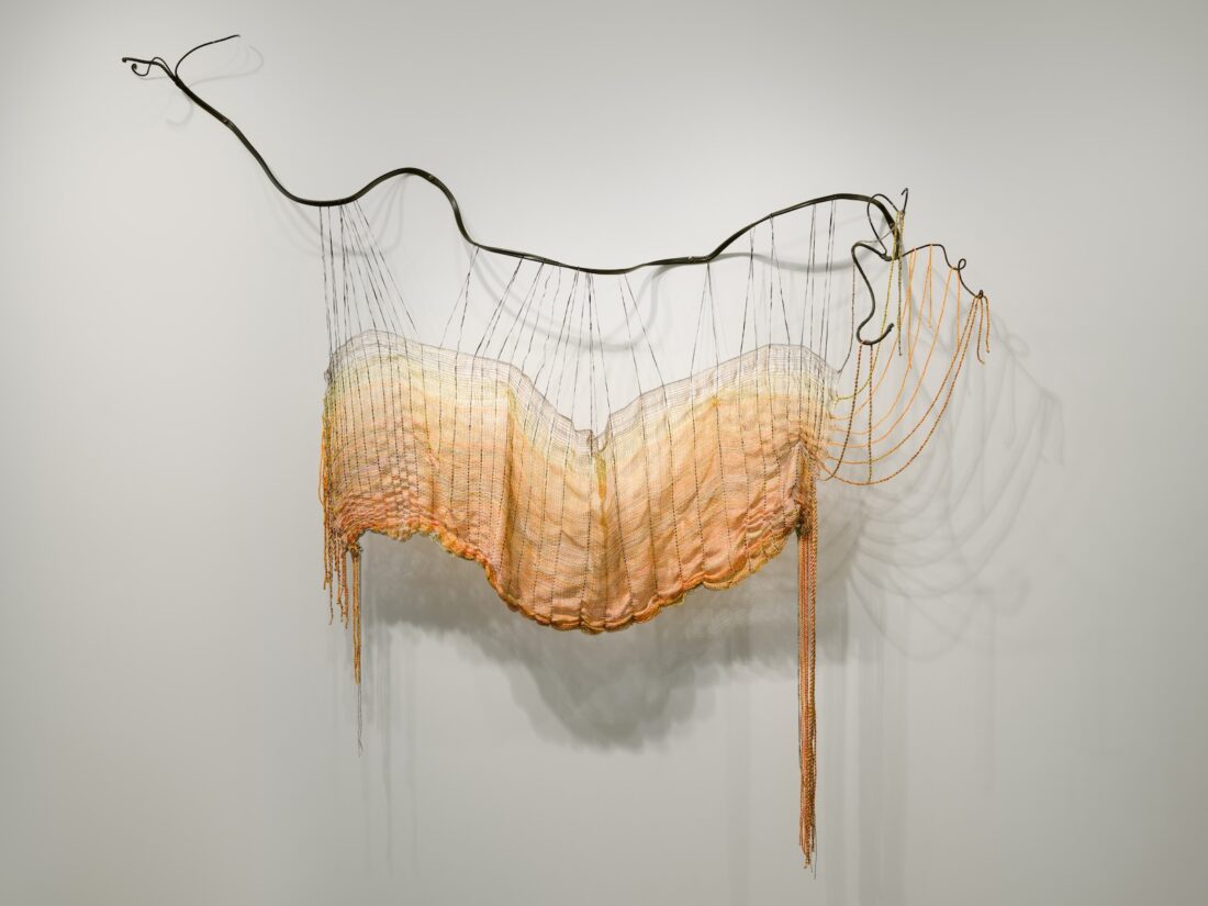 A wall-mounted artwork featuring woven, pastel-toned fibers draped and suspended from an undulating, dark metal rod, creating a cascading, organic shape with threads hanging loosely at the bottom and right side.