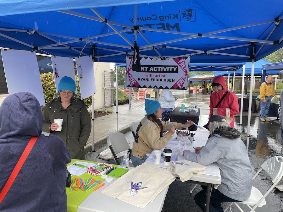 People gather under blue canopies at an outdoor art activity table on a rainy day. Some are seated and drawing, while others stand. Art supplies and tote bags are on the tables; a banner hangs overhead.